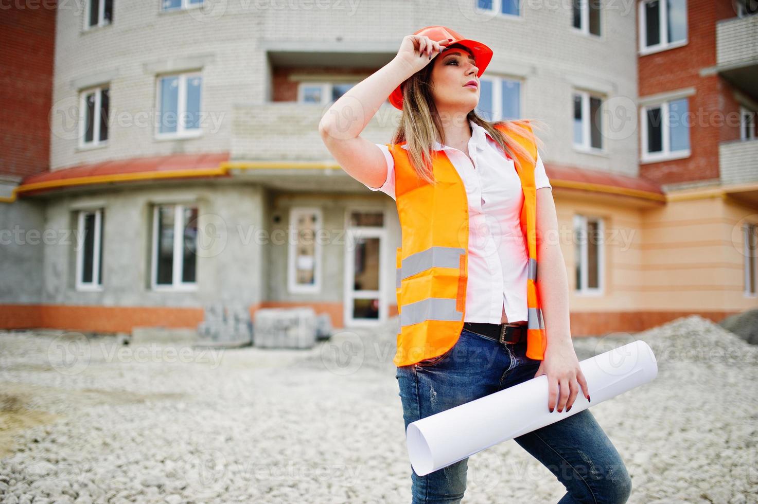 la donna del costruttore dell'ingegnere in gilet uniforme e casco protettivo arancione tiene la carta commerciale contro il nuovo edificio. tema del blocco abitativo della proprietà. foto