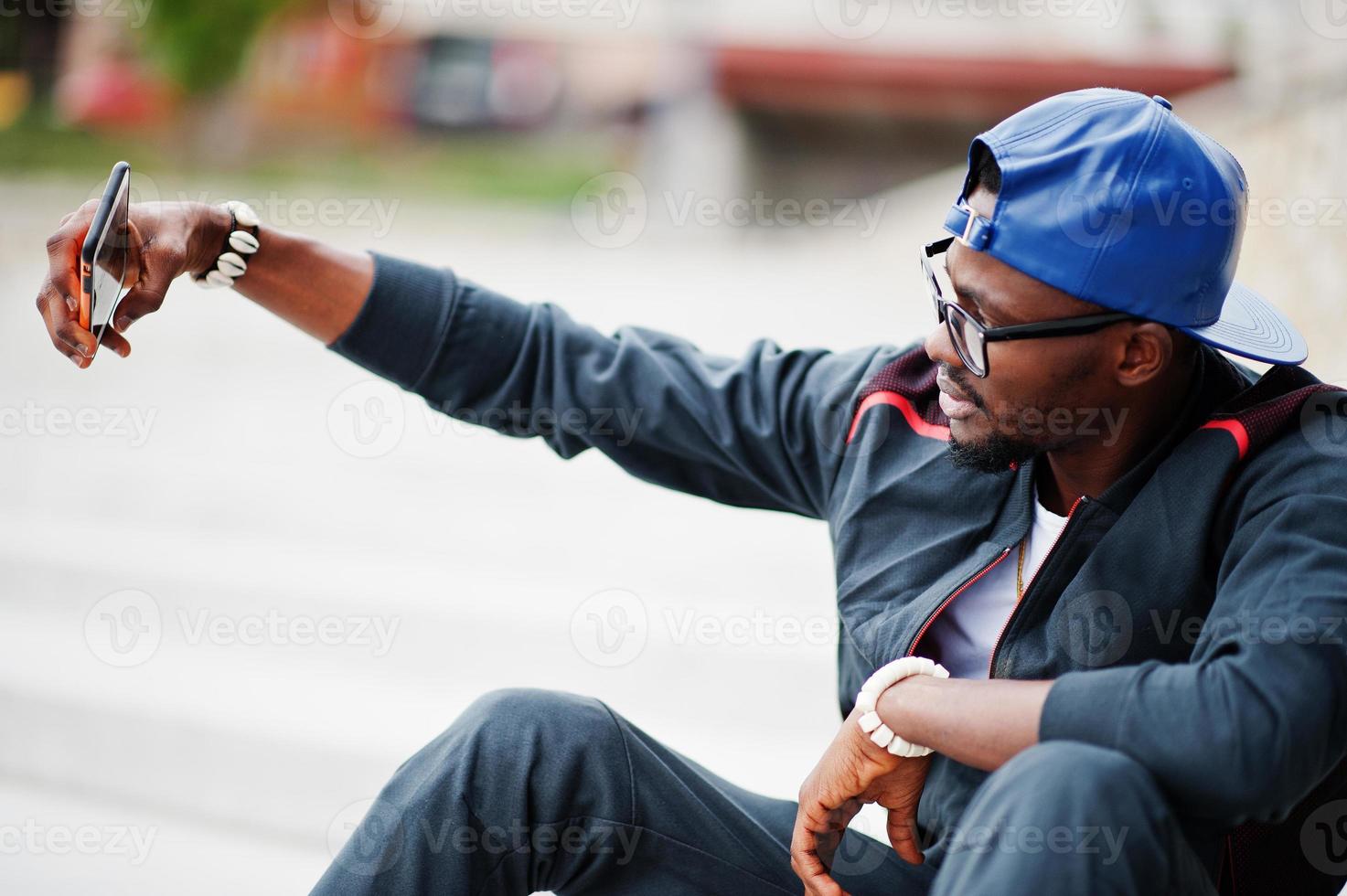 ritratto di elegante uomo afroamericano su abbigliamento sportivo, berretto e occhiali seduto sulle scale con il telefono a portata di mano, facendo selfie. uomini neri modello street fashion. foto