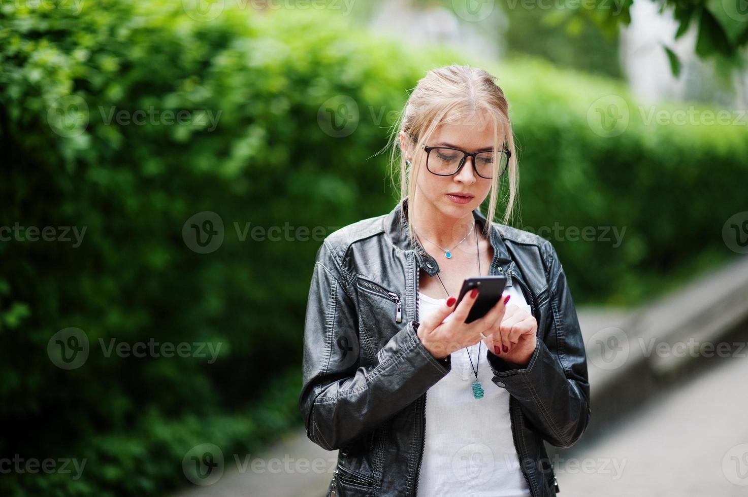 elegante donna bionda indossare jeans, occhiali e giacca di pelle con il telefono a portata di mano, contro i cespugli in strada. ritratto di modello urbano di moda. foto