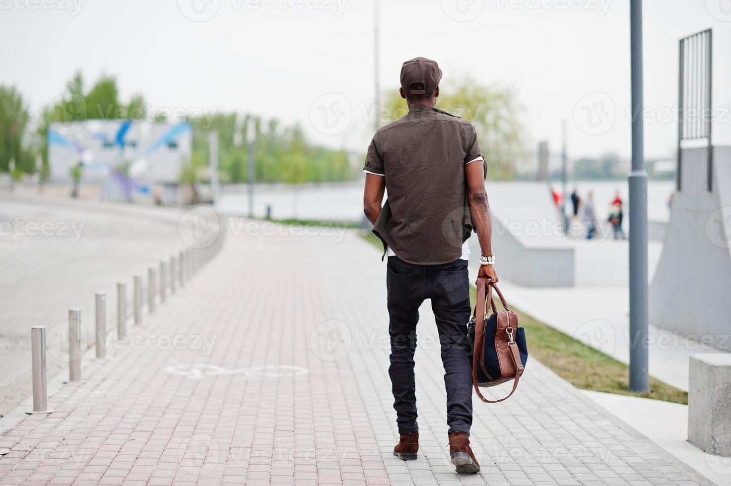 vista posteriore ritratto di camminare elegante uomo afroamericano indossare occhiali da sole e berretto con borsetta all'aperto. uomo nero di moda di strada. foto
