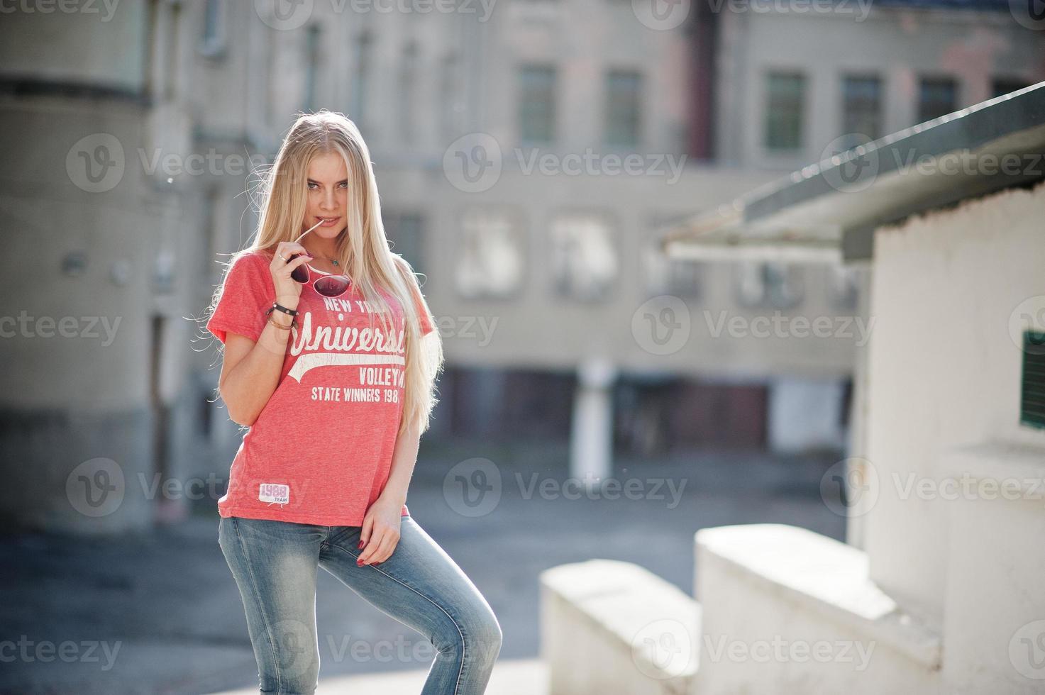 elegante donna bionda felice indossare jeans, occhiali da sole e t-shirt poste in strada con tempo soleggiato. ritratto di modello urbano di moda. foto