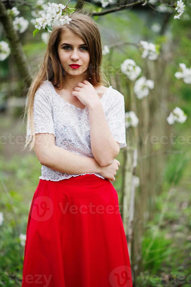 ritratto di bella ragazza con labbra rosse al giardino di fiori primaverili, indossare su abito rosso e camicetta bianca. foto