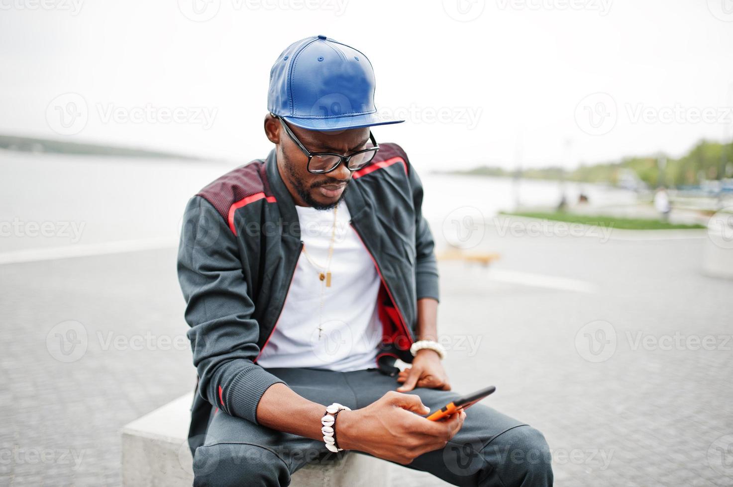 ritratto di elegante uomo afroamericano su abbigliamento sportivo, berretto e occhiali seduto al cubo di pietra con il telefono a portata di mano. uomini neri modello street fashion. foto