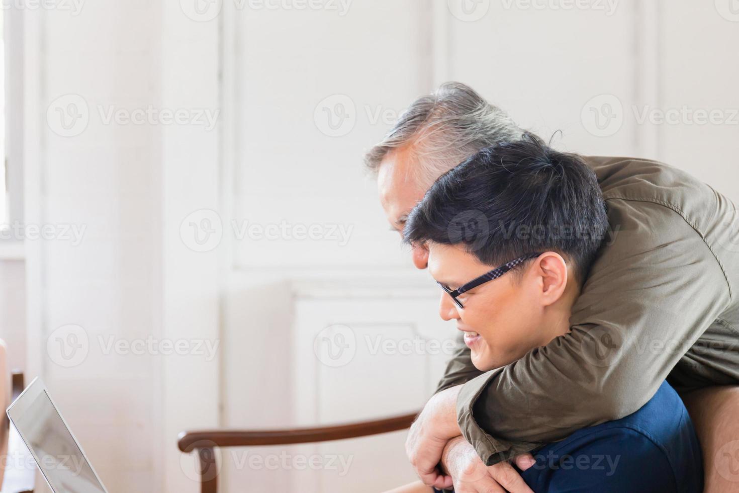 felice padre asiatico senior e figlio adulto utilizzando computer portatile parlando in videochiamata in soggiorno foto