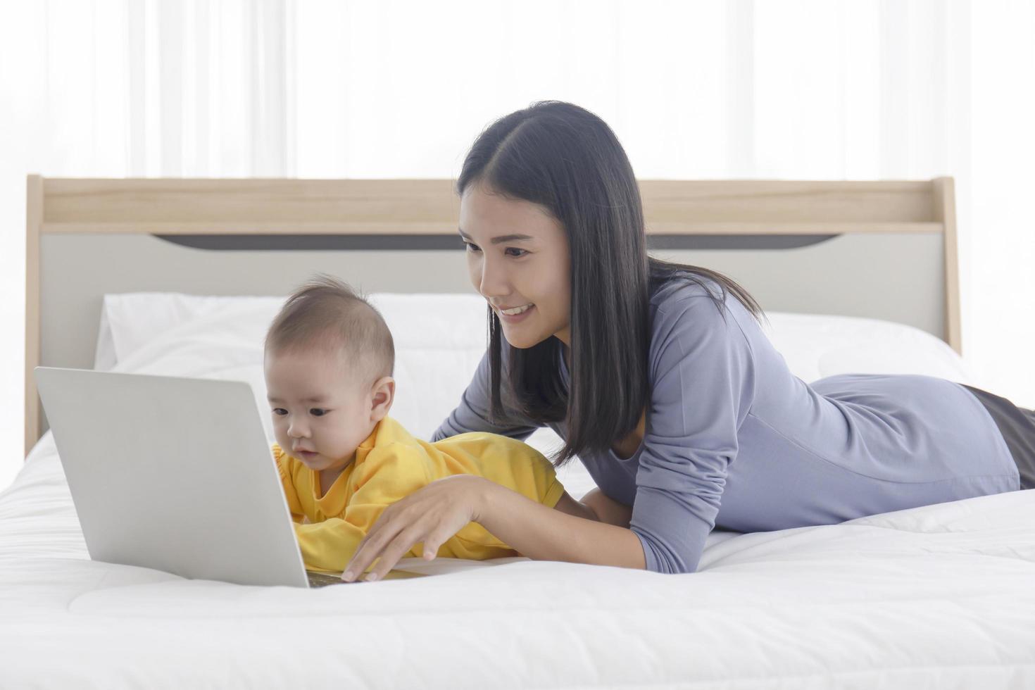 una mamma asiatica sta lavorando su un laptop a letto e con il suo bambino accanto al concetto di lavoro a casa. foto