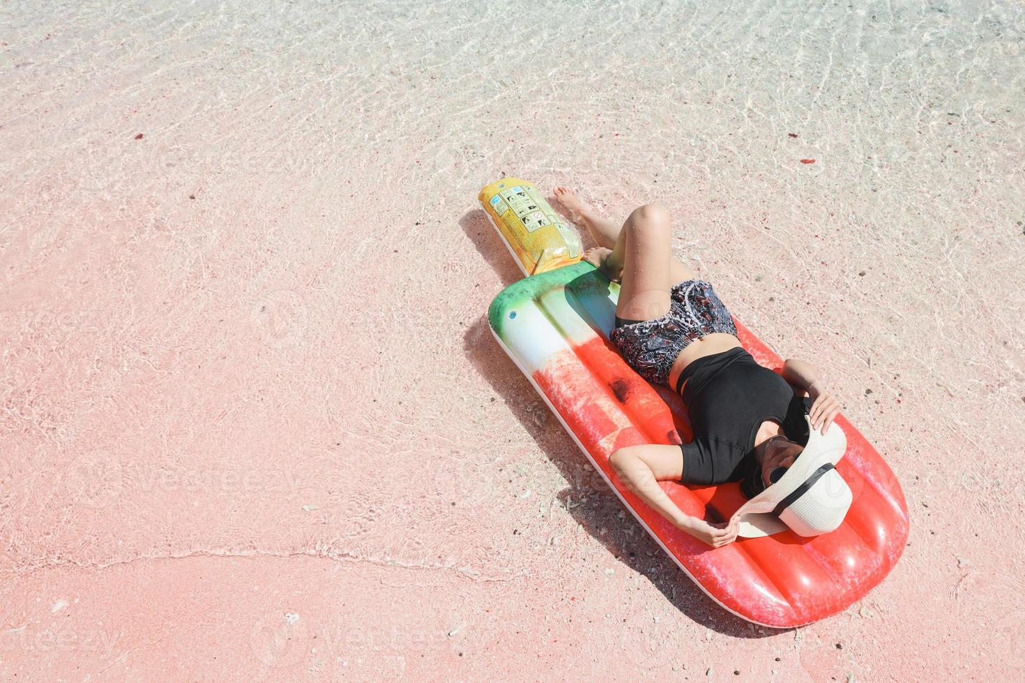 una ragazza in cappello estivo che prende il sole sul materasso gonfiabile in spiaggia di sabbia rosa foto