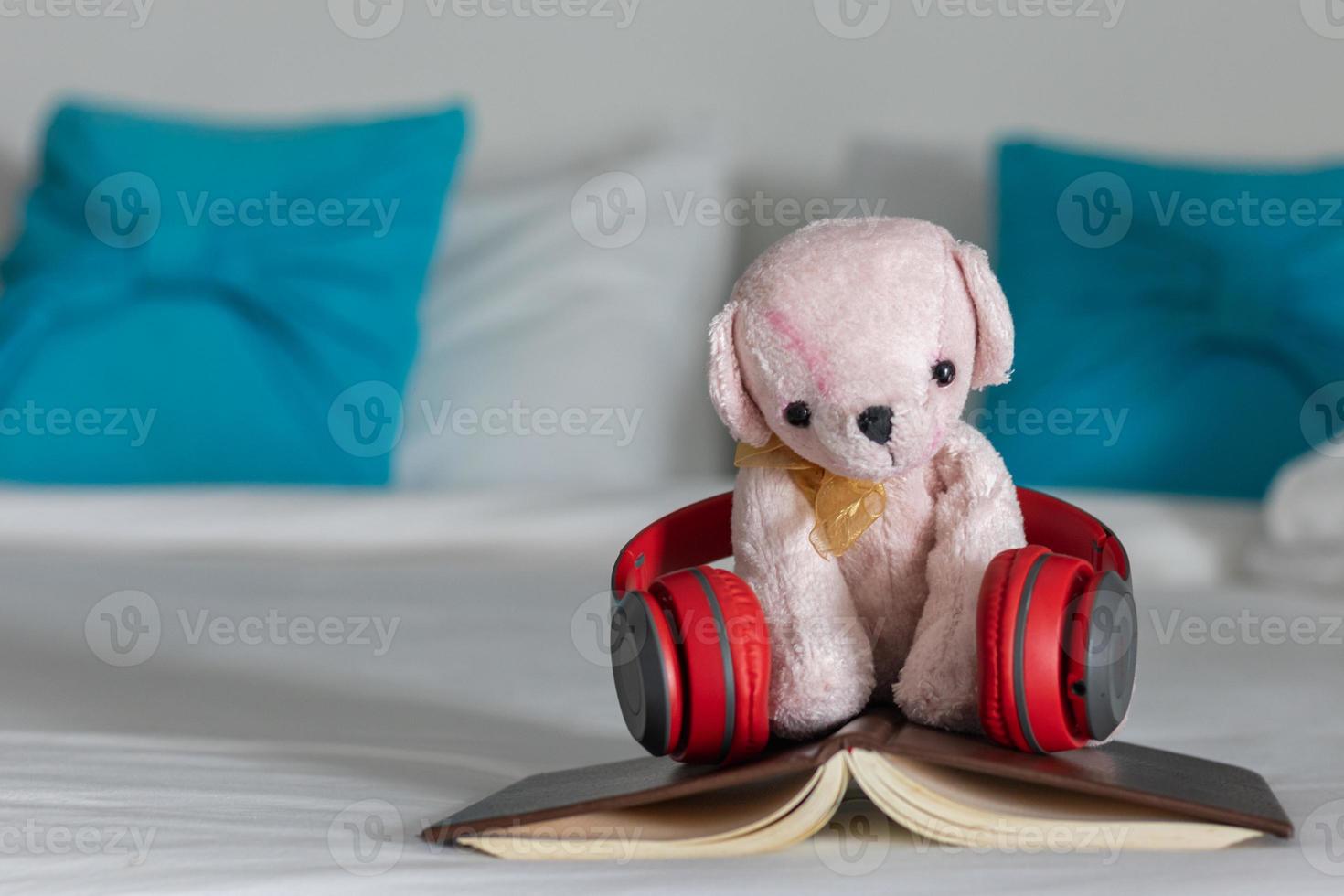 una cuffia rossa è posizionata su un libro vicino a un orsacchiotto rosa sul letto al mattino, pronto per un comodo riposo. un paio di cuffie e un libro poggiano sulla biancheria bianca in camera da letto. foto