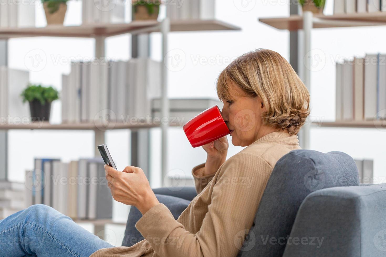 donna rilassante con tazze di caffè in soggiorno, donna matura che utilizza smartphone mobile durante una pausa caffè foto