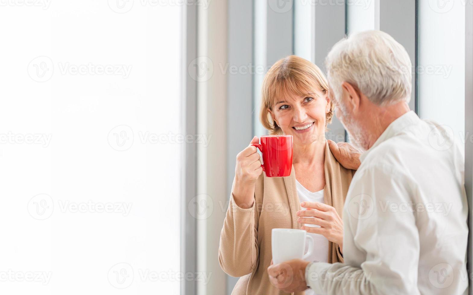 coppia di anziani che parla stando in piedi vicino alla finestra con tazze di caffè, coppia senior all'interno della nuova casa durante la pausa caffè foto