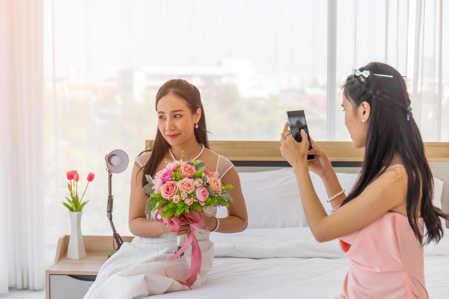 l'amica della sposa sta usando uno smartphone per scattare una foto di una sposa asiatica in un abito da sposa in mano, con in mano un grande e bellissimo mazzo di fiori seduto sul letto.