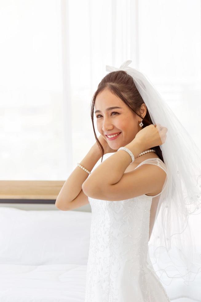 una sposa asiatica vestita con un abito da sposa bianco sta in un sorriso carino e luminoso accanto al letto nella stanza. foto
