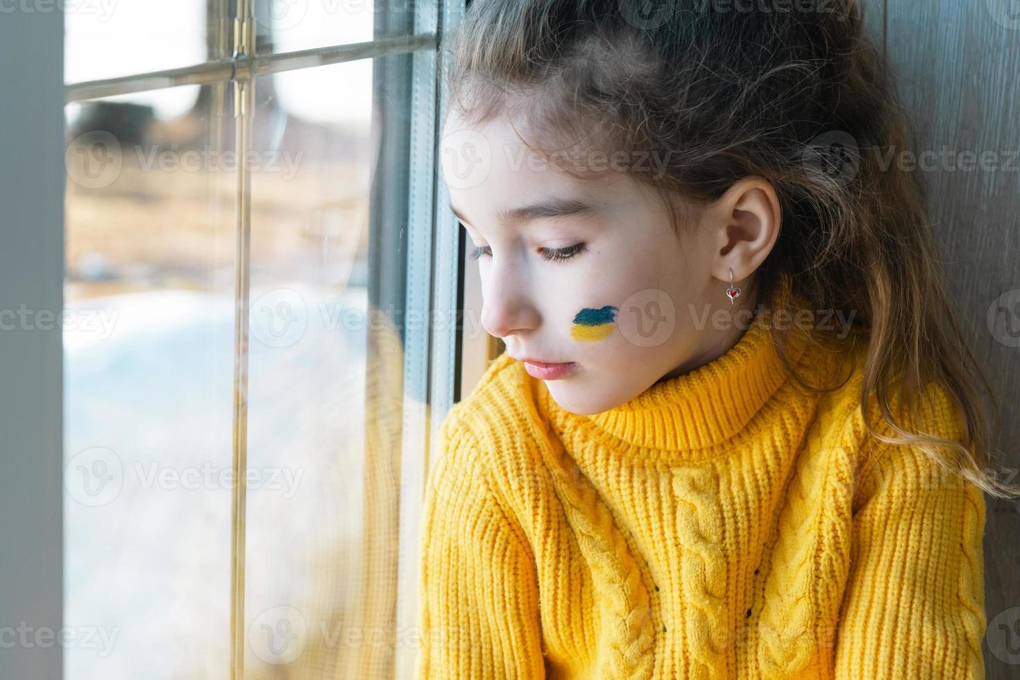 un bambino triste guarda la finestra con la bandiera dell'ucraina dipinta sulla guancia, preoccupazioni e paure. aiuti umanitari ai bambini, pace nel mondo, sicurezza. foto