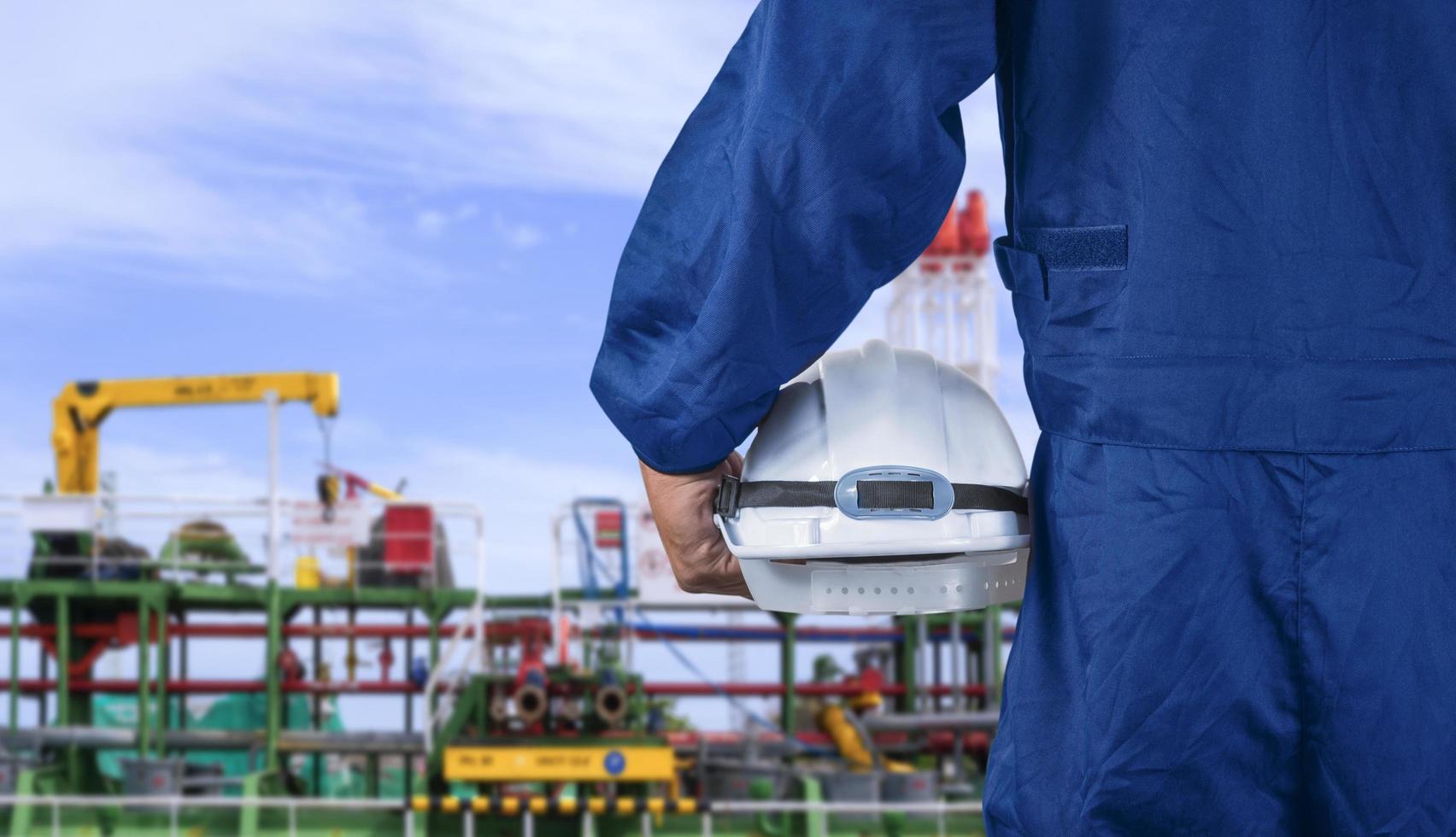 primo piano la mano dell'ingegnere nella tuta blu del meccanico che tiene il casco di sicurezza bianco prima di lavorare su una petroliera grezza nell'area del cantiere navale foto