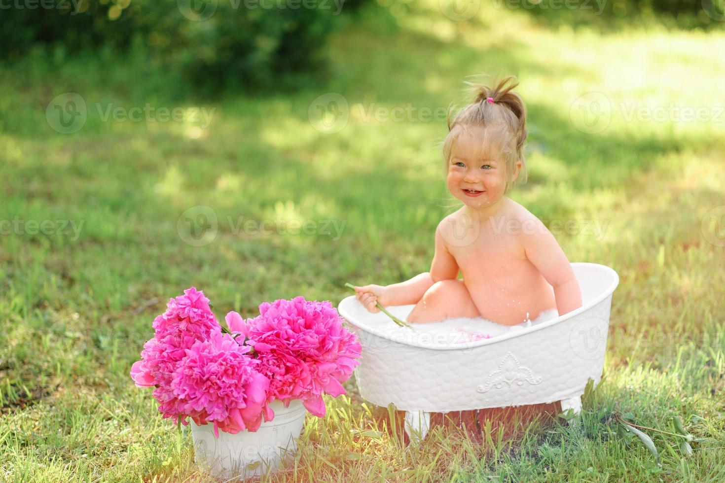 la ragazza felice del bambino prende un bagno di latte con i petali. bambina in un bagno di latte su sfondo verde. mazzi di peonie rosa. fare il bagnetto. igiene e cura dei bambini piccoli. foto