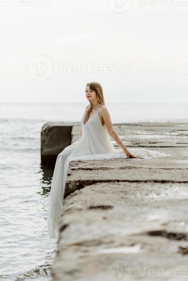 una giovane bella ragazza con un lungo vestito color latte cammina lungo la spiaggia e il molo sullo sfondo del mare. foto