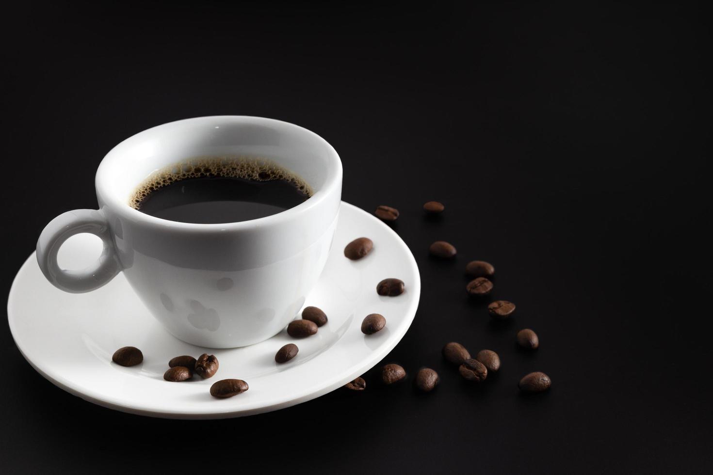 caffè nero in bianco tazza di caffè e piattino con caffè in grani su sfondo nero. foto