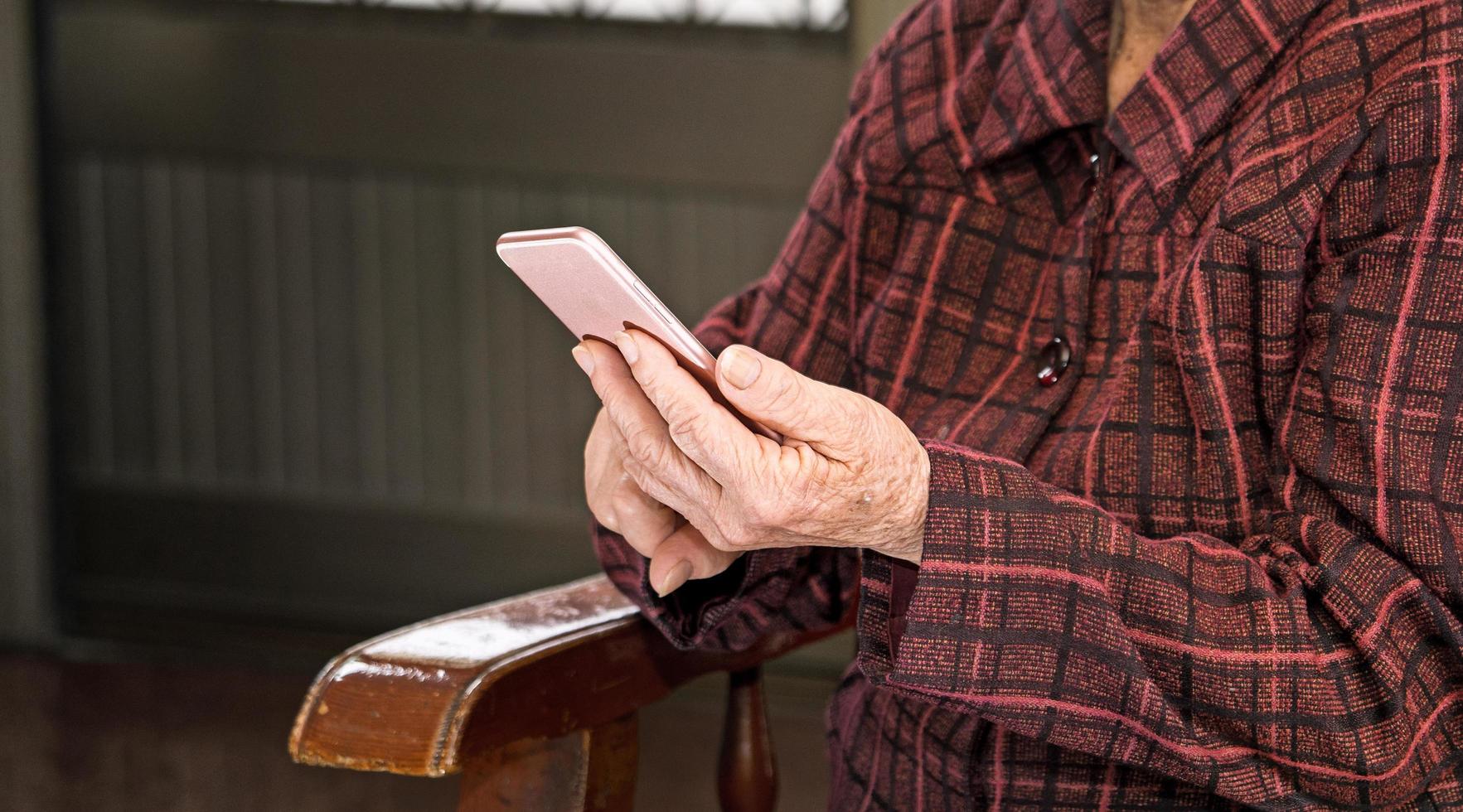 donna anziana asiatica seduta e guardando attraverso qualcosa su uno smartphone moderno, collegandosi con gli altri a casa, tecnologia vivente, primo piano foto