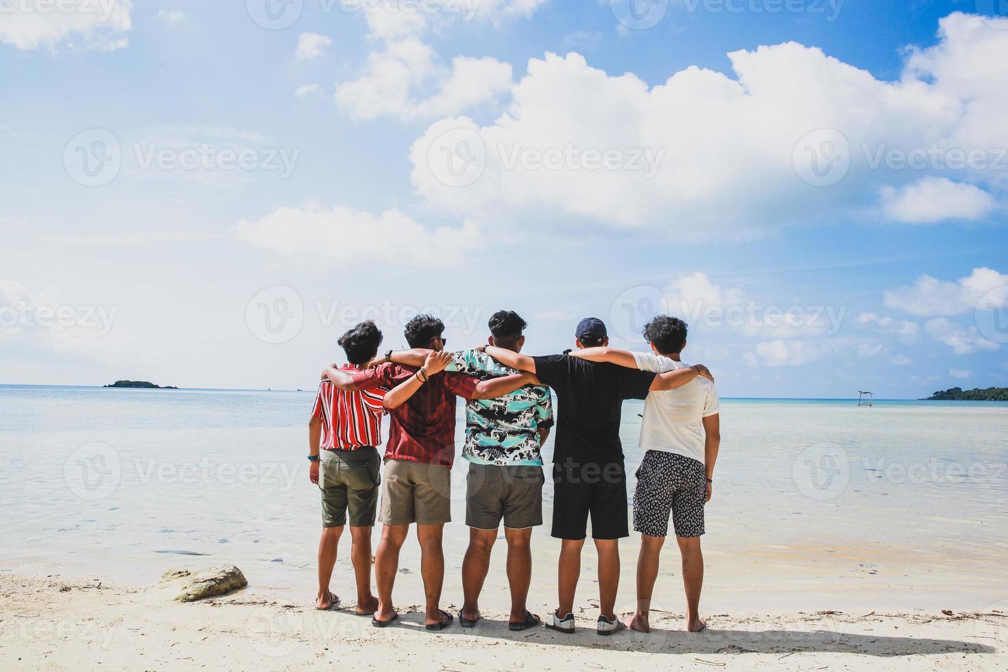 vista posteriore di un gruppo di amici in piedi e abbracciarsi sulla spiaggia tropicale durante le vacanze foto