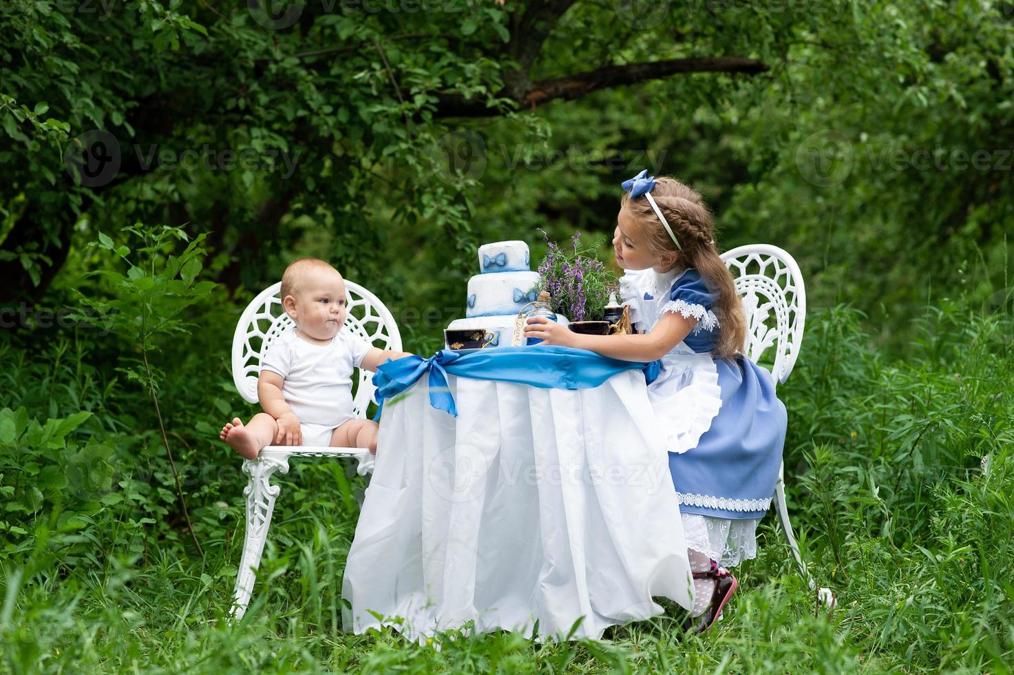 una ragazzina carina con il costume Alice del Paese delle Meraviglie e suo fratello di un anno stanno organizzando un tea party al loro tavolo magico. fotografato in natura. foto