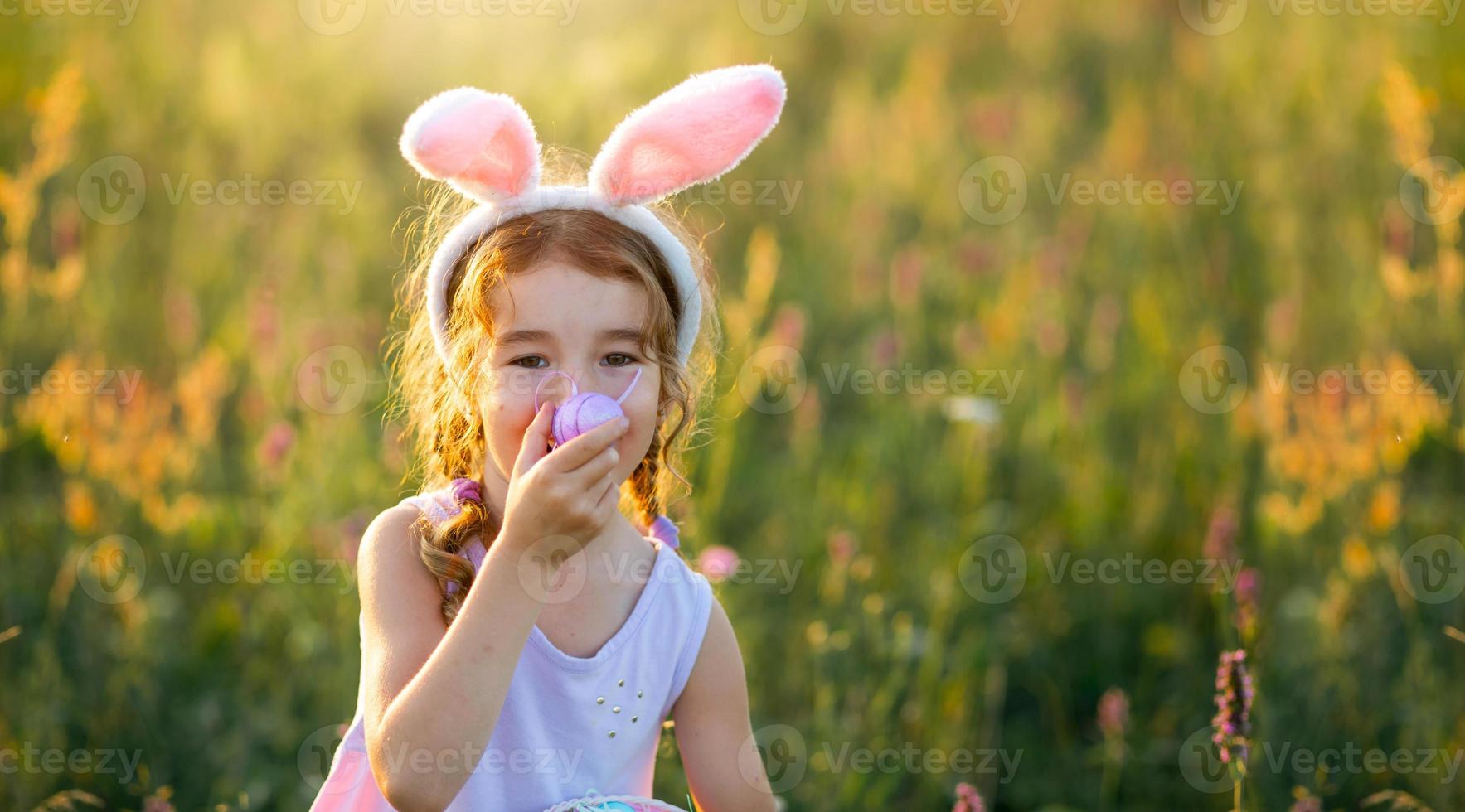 ragazza carina e divertente con uova di Pasqua dipinte in primavera in natura in un campo con luce solare dorata e fiori. vacanze di pasqua, coniglietto pasquale con le orecchie, uova colorate in un cestino. stile di vita foto