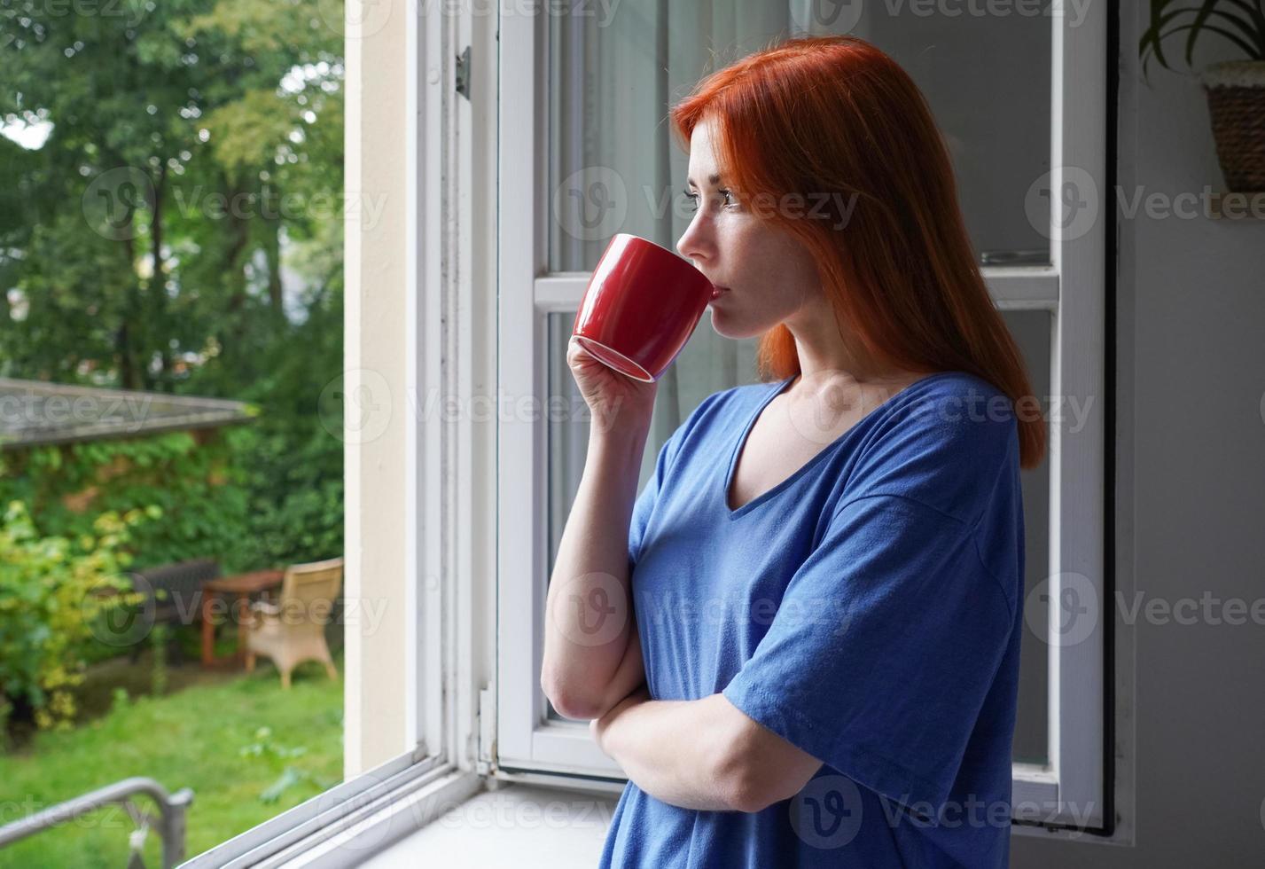 giovane donna che guarda fuori dalla finestra mentre beve dalla tazza di caffè foto