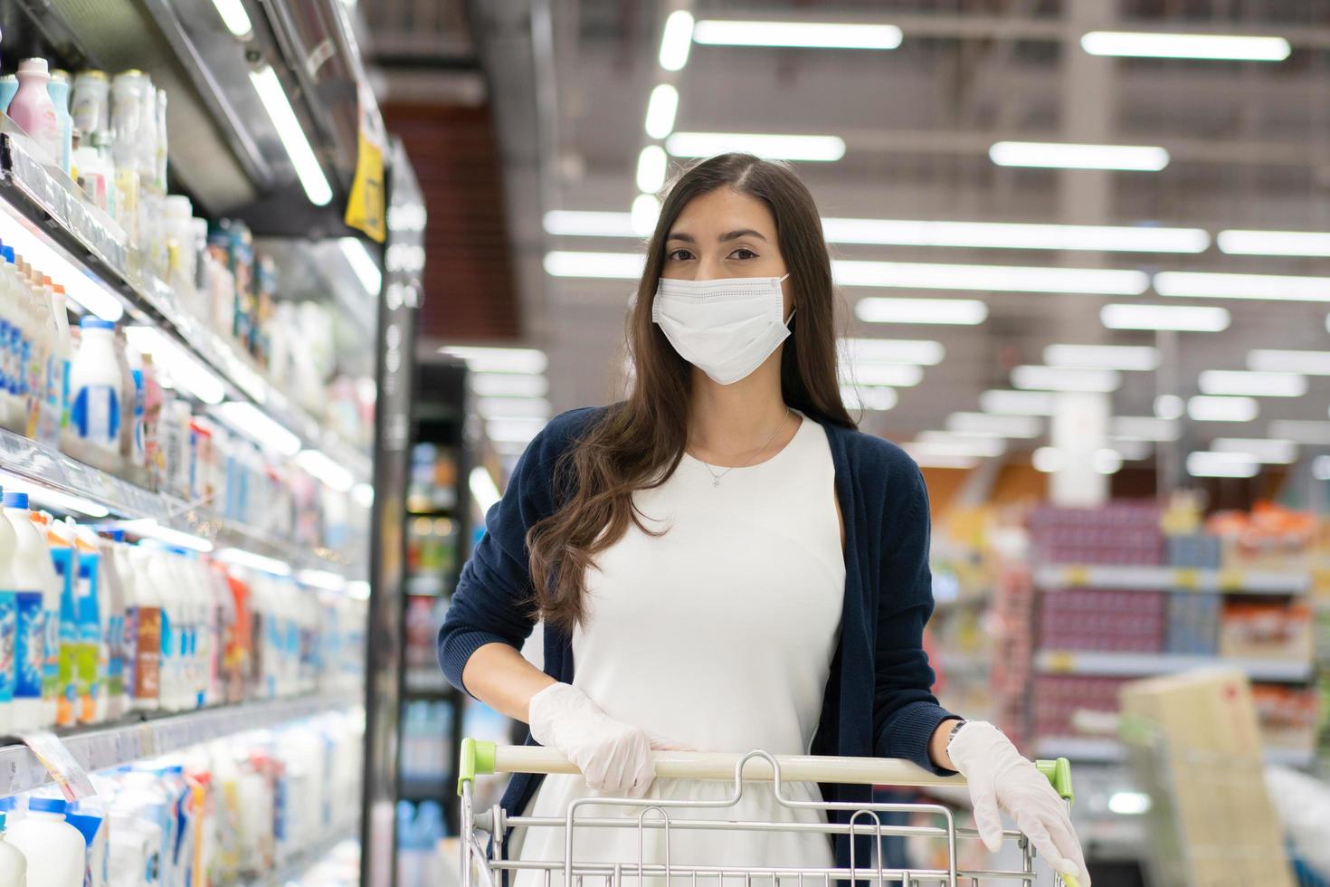 donna che indossa una maschera medica e un guanto di gomma spinge il carrello della spesa nel grande magazzino del supermercato. ragazza che guarda lo scaffale della drogheria. nuovo stile di vita normale durante il coronavirus o la pandemia covid foto