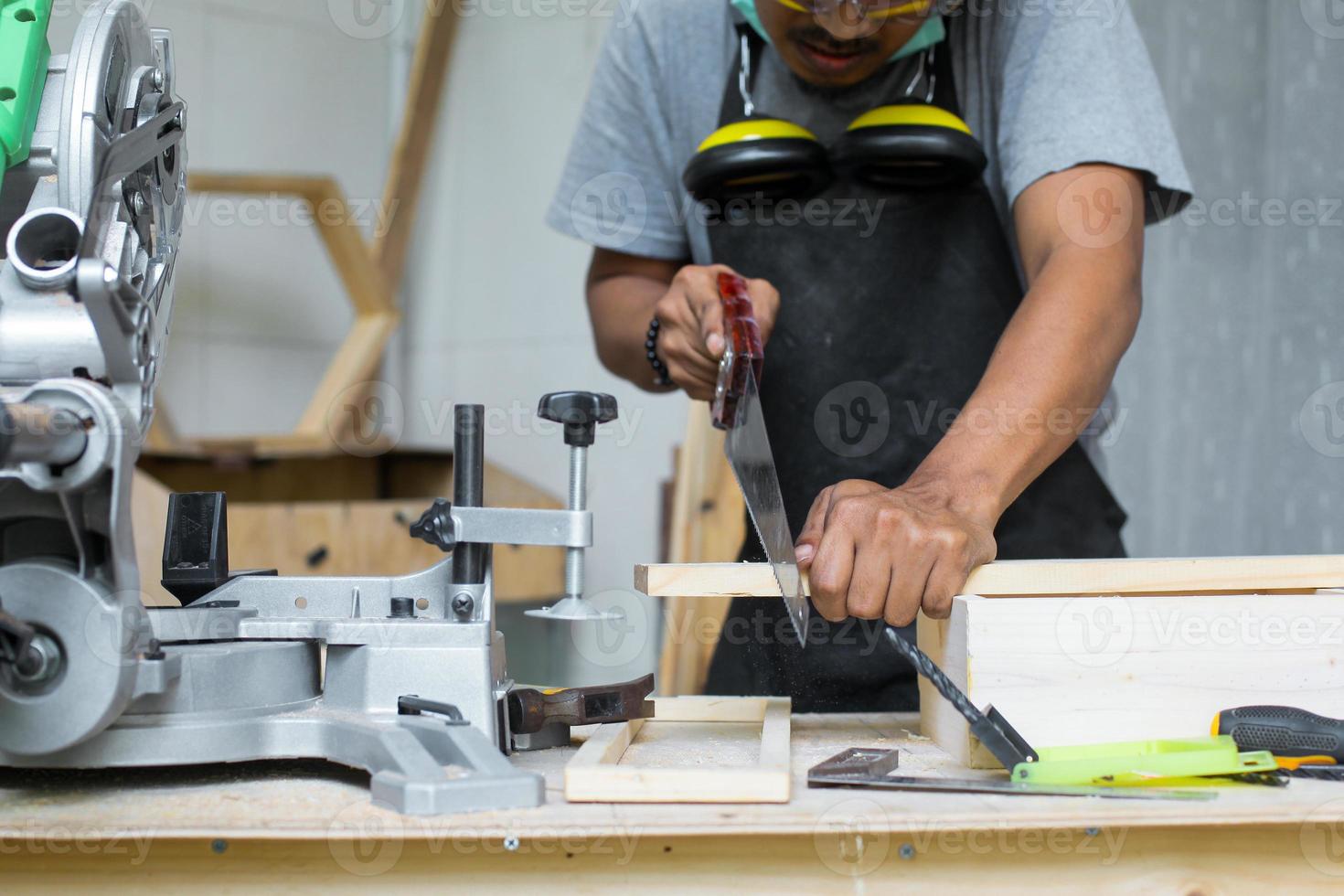 un giovane falegname maschio che taglia il legno usando una sega a mano sul suo tavolo da officina indossando attrezzature di sicurezza foto