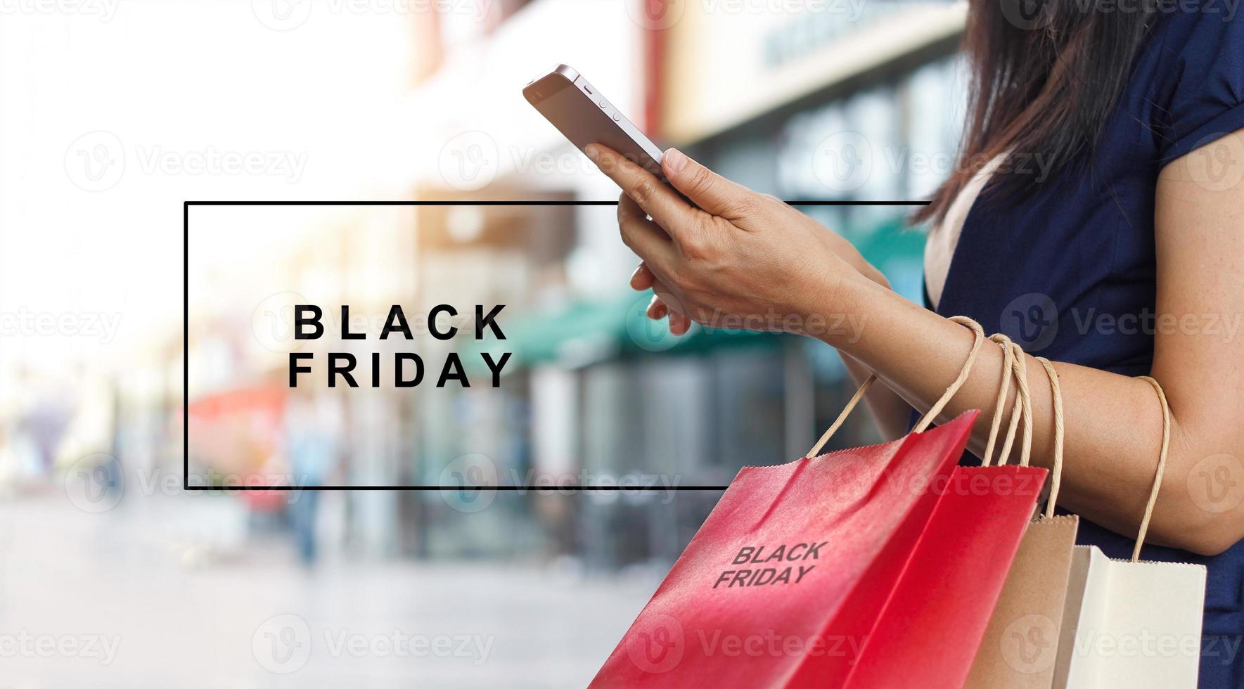 venerdì nero, donna che usa lo smartphone e tiene in mano la borsa della spesa mentre si trova sullo sfondo del centro commerciale foto