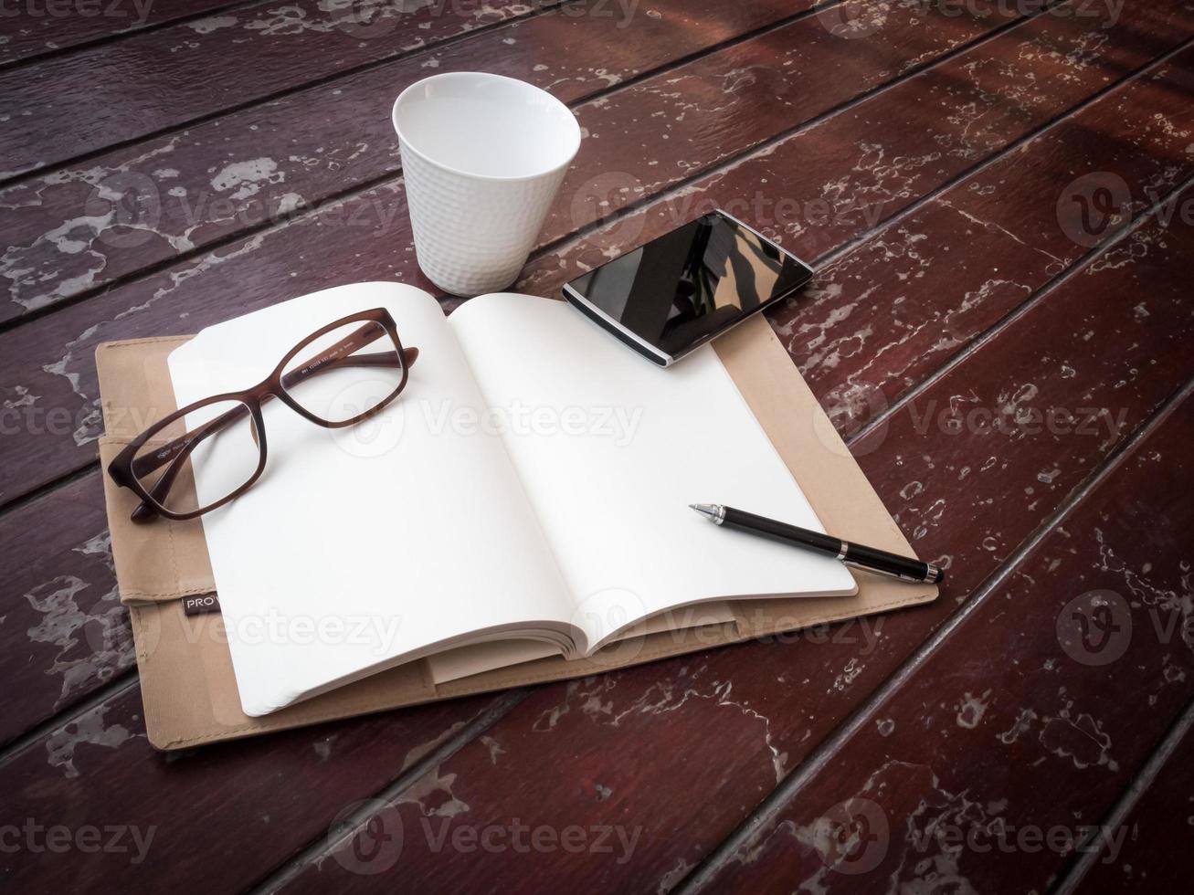 area di lavoro con occhiali, penna, smartphone e tazza di caffè, carta per appunti e taccuino sul vecchio tavolo di legno foto