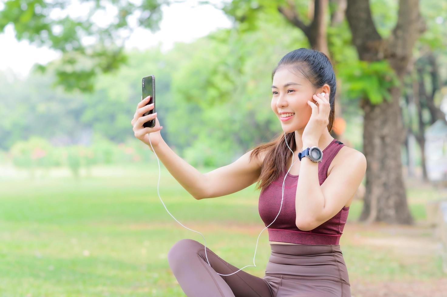 una bella donna asiatica si prende una pausa per ascoltare la musica sul suo cellulare dopo aver fatto esercizio nel parco foto