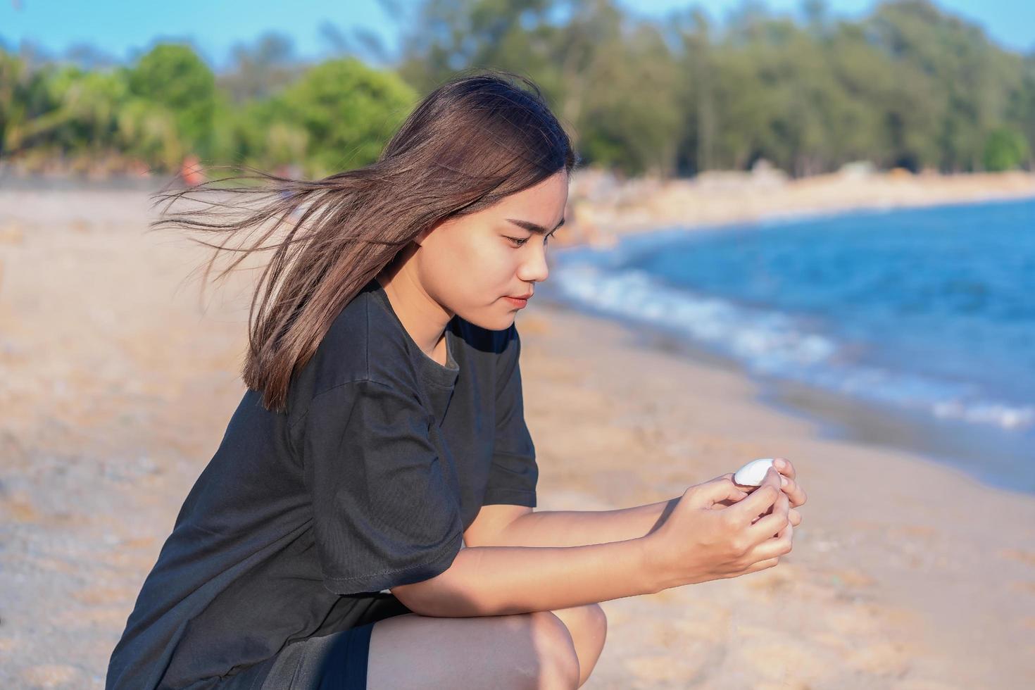 ragazza adolescente asiatica carina pensando da sola e guardando il guscio bianco seduto sulla sabbia della spiaggia foto