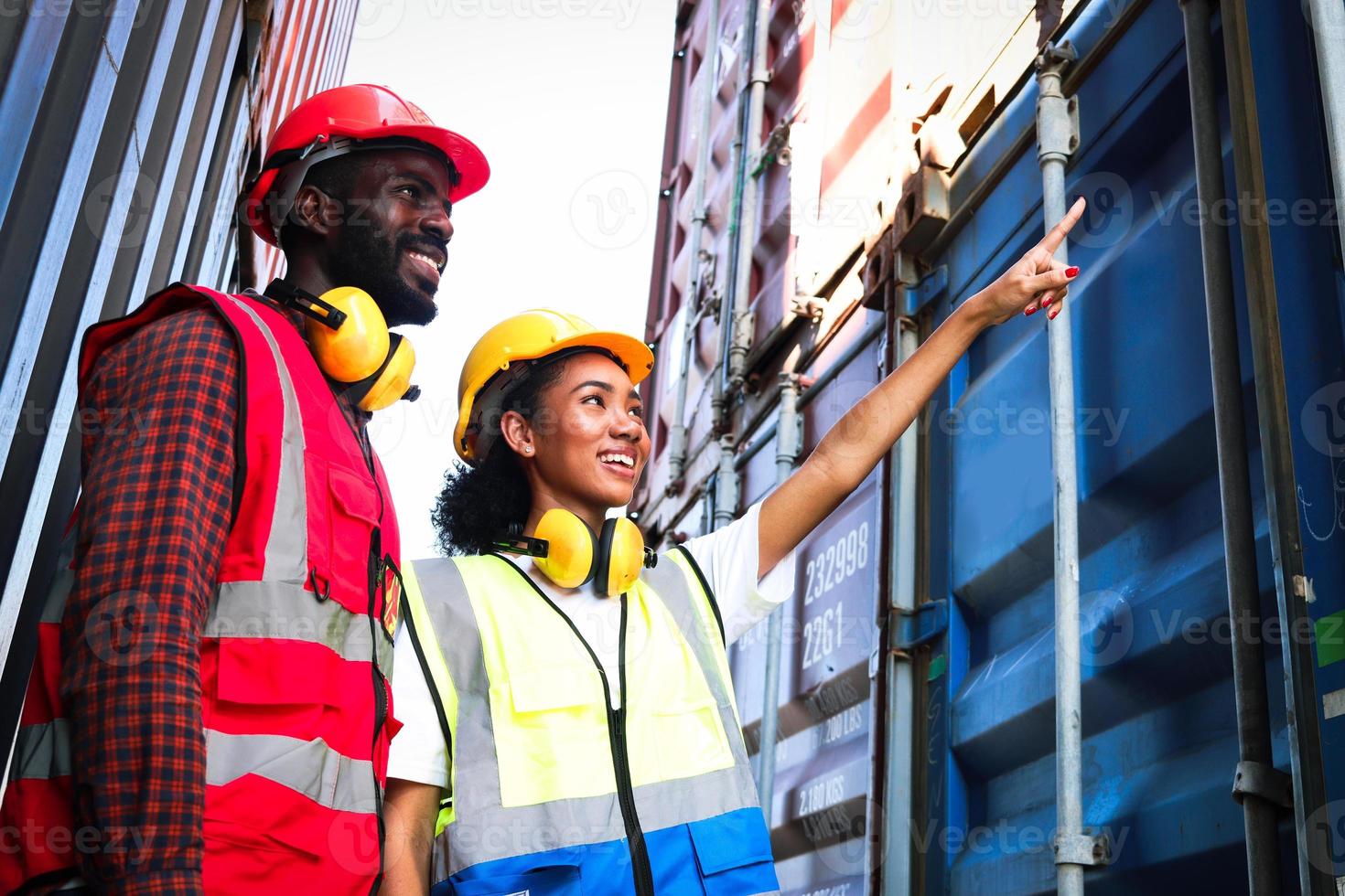 due ingegneri afroamericani industriali, uomini e donne, indossano giubbotto di sicurezza e casco che lavorano insieme al cantiere logistico di container per carichi di spedizione, lavoratrice che punta e chiede parere al collega. foto