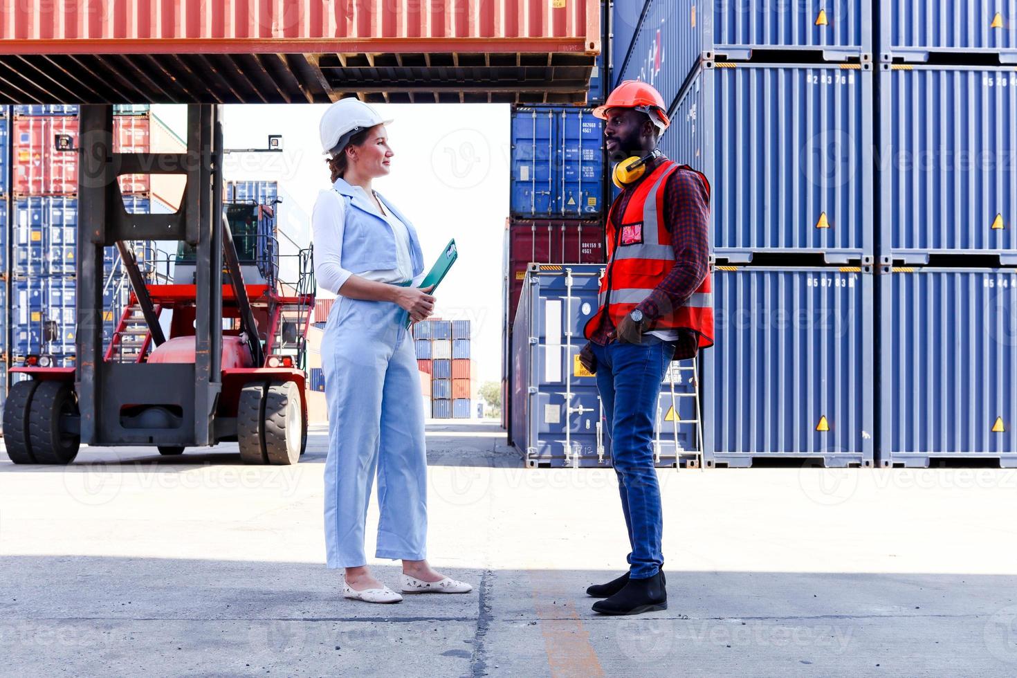 due lavoratori che indossano un giubbotto di sicurezza e un casco che discutono al cantiere di container per carichi logistici. uomo dell'ingegnere afroamericano che parla con il capo della bella giovane donna con i capelli biondi sul posto di lavoro. foto