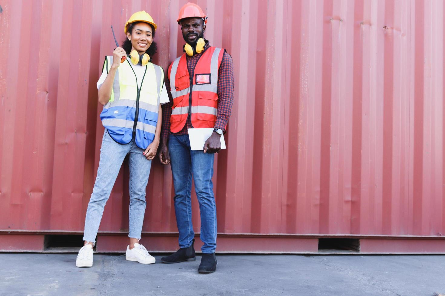 Ritratto di due uomini e donne ingegnere afroamericano industriale sorridenti felici che indossano giubbotto di sicurezza e casco in piedi davanti al container al cantiere di carico logistico. foto