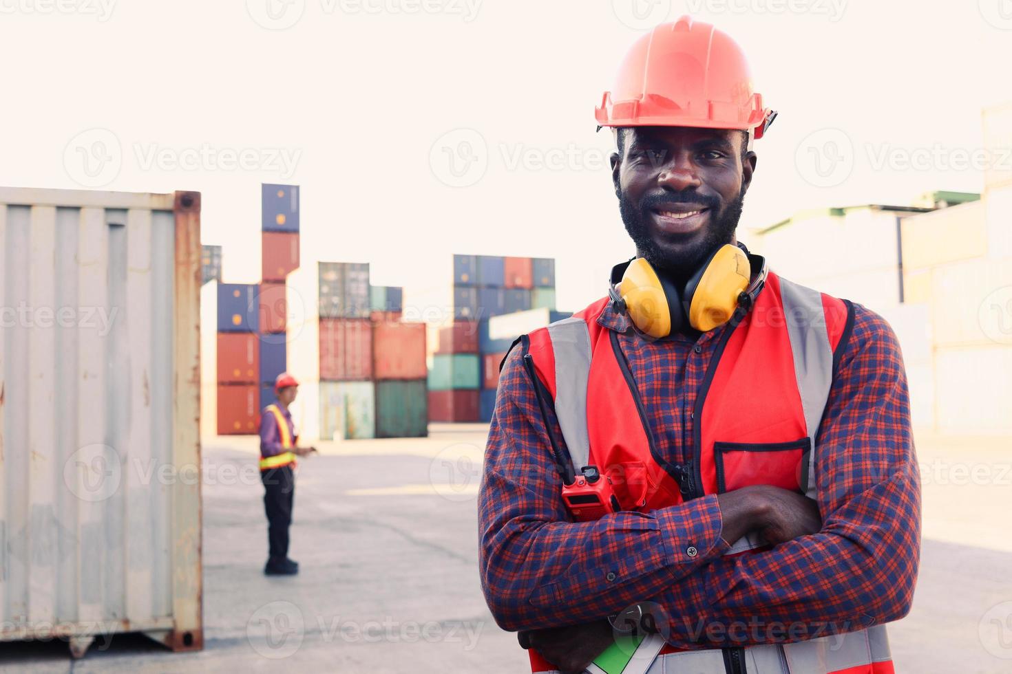 ritratto di felice sorridente africano americano giovane ingegnere lavoratore uomo che indossa sicurezza luminoso colore rosso neon giubbotto e casco, in piedi con le braccia incrociate al deposito di container di carico logistico foto