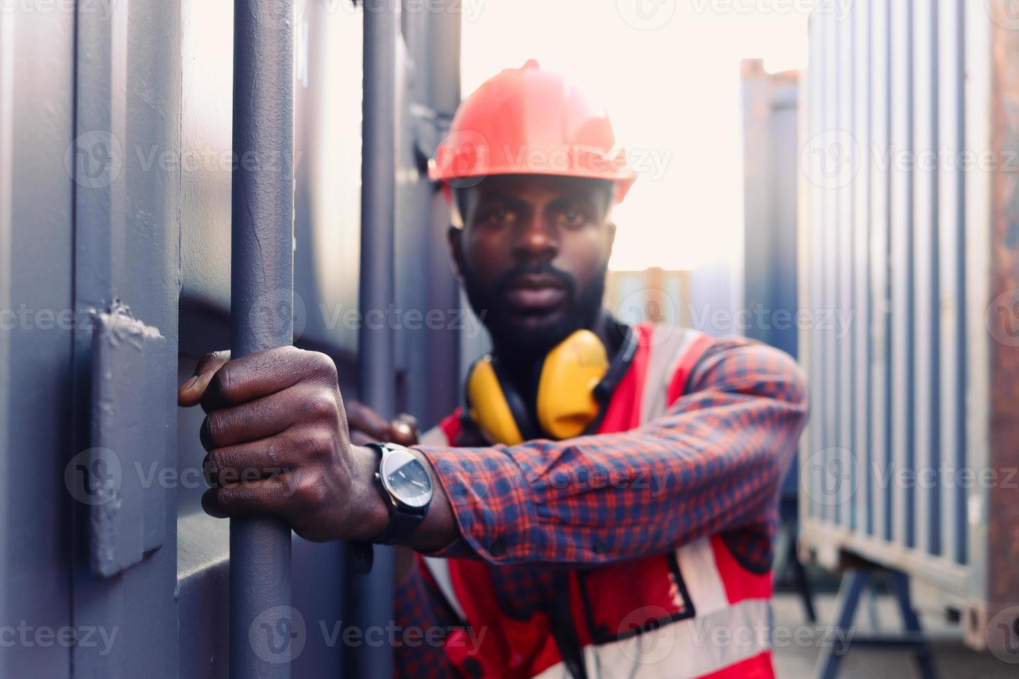 Ritratto di giovane ingegnere afroamericano lavoratore uomo che indossa un giubbotto e casco di sicurezza di colore rosso neon luminoso, cercando di aprire una porta del container di spedizione presso il cantiere di container logistici. foto