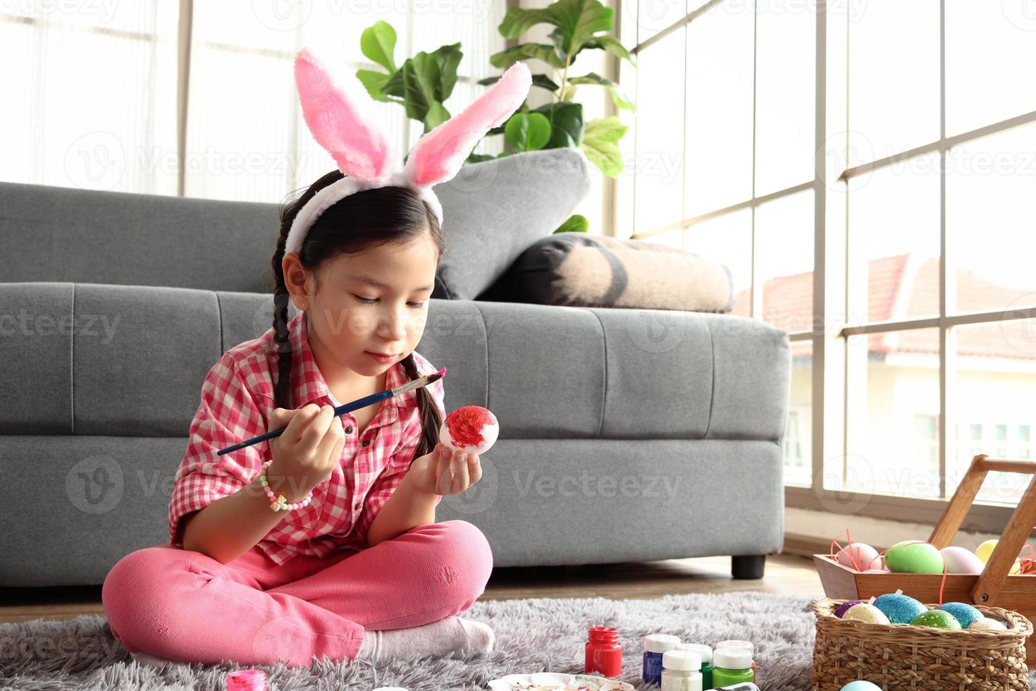 adorabile rosa coniglietta pasquale bambina con orecchie di coniglio fascia che dipinge uova di Pasqua colorate in soggiorno, decorazioni per celebrare l'inizio della primavera. foto