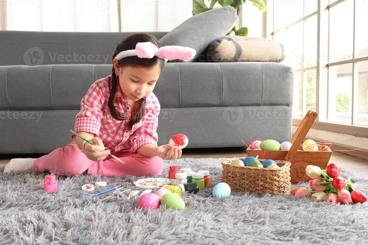 adorabile rosa coniglietta pasquale bambina con orecchie di coniglio fascia che dipinge uova di Pasqua colorate in soggiorno, decorazioni per celebrare l'inizio della primavera. foto
