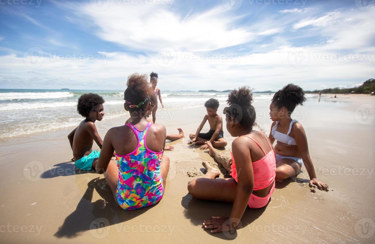 bambini che giocano a correre sulla sabbia in spiaggia, un gruppo di bambini che si tengono per mano in fila sulla spiaggia in estate, vista posteriore contro mare e cielo blu foto