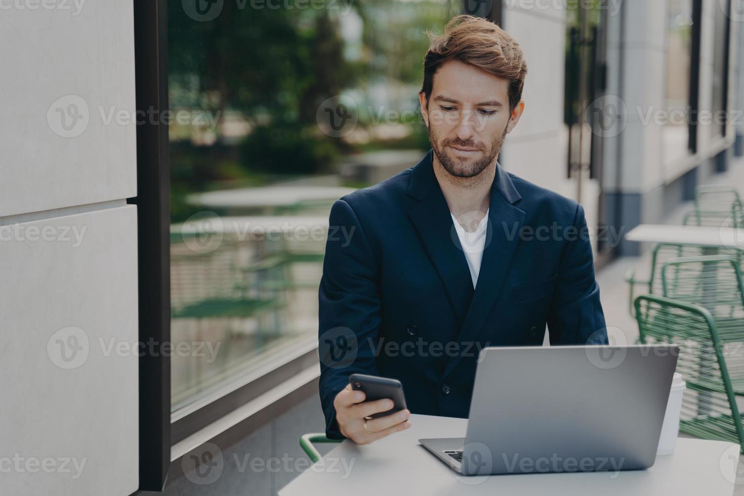 il capo maschio serio vestito con abiti eleganti lavora online su un computer portatile con cellulare foto