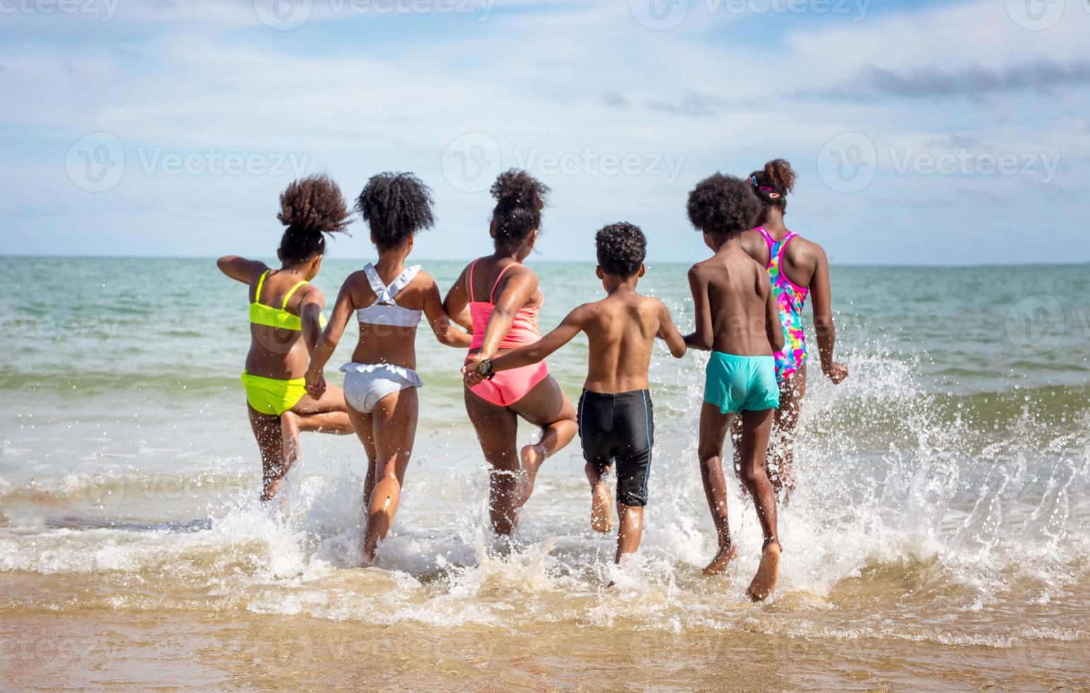 bambini che giocano a correre sulla sabbia in spiaggia, un gruppo di bambini che si tengono per mano in fila sulla spiaggia in estate, vista posteriore contro mare e cielo blu foto