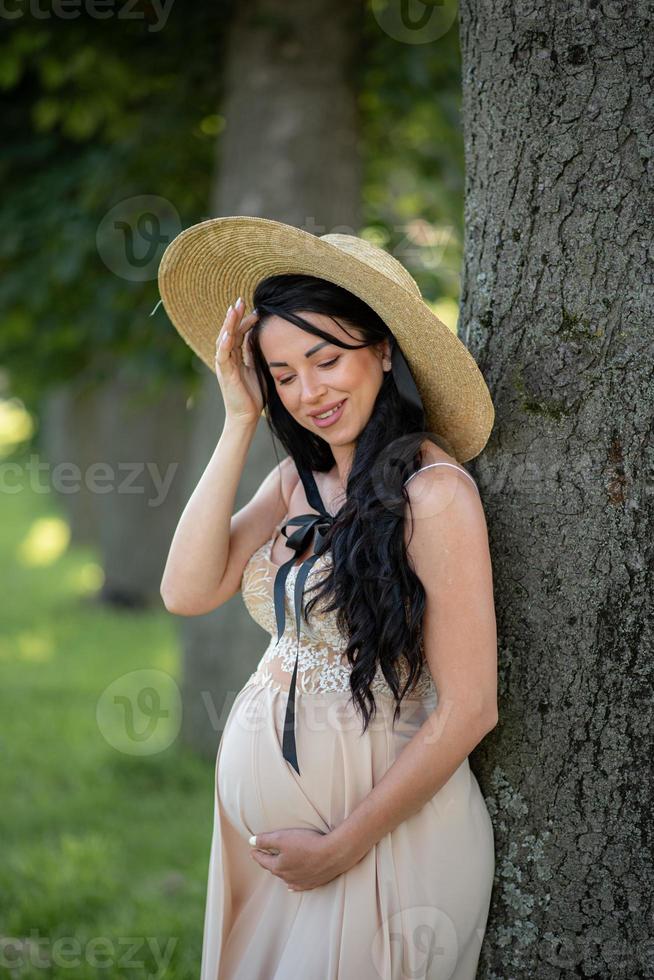 donna incinta in un cappello che posa in un vestito su uno sfondo di alberi verdi. foto