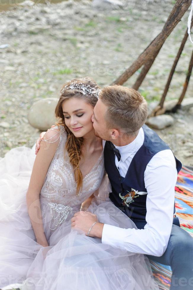 una coppia di sposini attraenti, un momento felice e gioioso. un uomo e una donna si radono e si baciano in abiti da festa. cerimonia nuziale in stile bohémien nella foresta all'aria aperta. foto