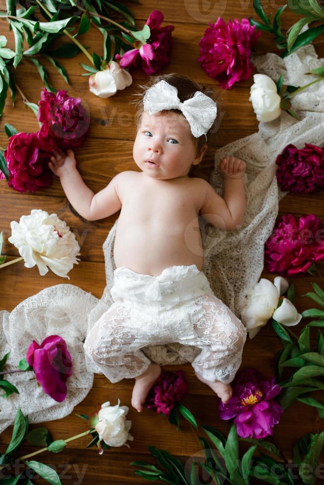 una bambina di due mesi giace su un tavolo con delle peonie. foto