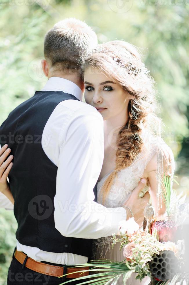 coppia attraente sposi novelli, momento felice e gioioso. l'uomo e la donna in abiti festivi si siedono sulle pietre vicino alla decorazione del matrimonio in stile boho. cerimonia all'aperto. foto