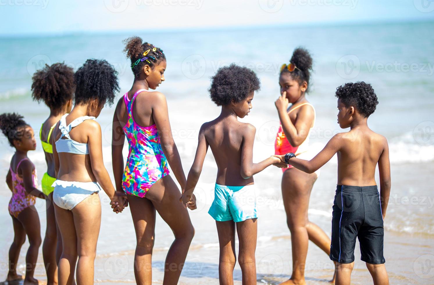 bambini che giocano a correre sulla sabbia in spiaggia, un gruppo di bambini che si tengono per mano in fila sulla spiaggia in estate, vista posteriore contro mare e cielo blu foto