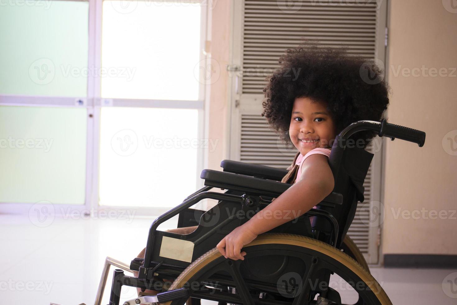 bambina di etnia afro-americana che riposa sulla sedia a rotelle in attesa di vedere il medico in ospedale. ragazza afro-americana seduta su una sedia a rotelle e sorridente alla telecamera. foto