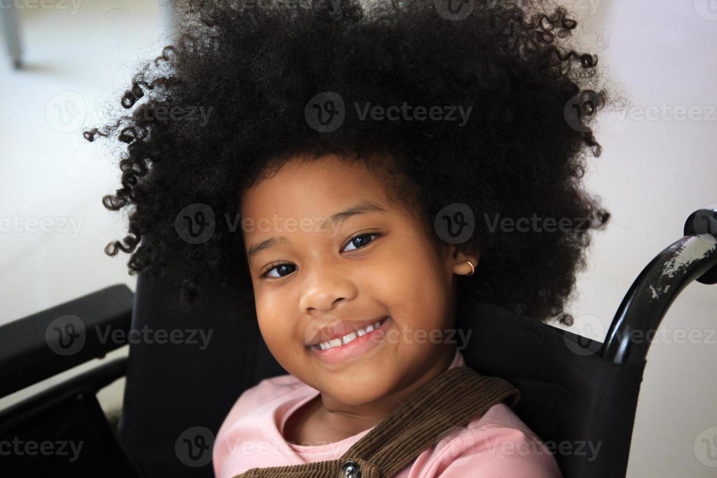 bambina di etnia afro-americana che riposa sulla sedia a rotelle in attesa di vedere il medico in ospedale. ragazza afro-americana seduta su una sedia a rotelle e sorridente alla telecamera. foto