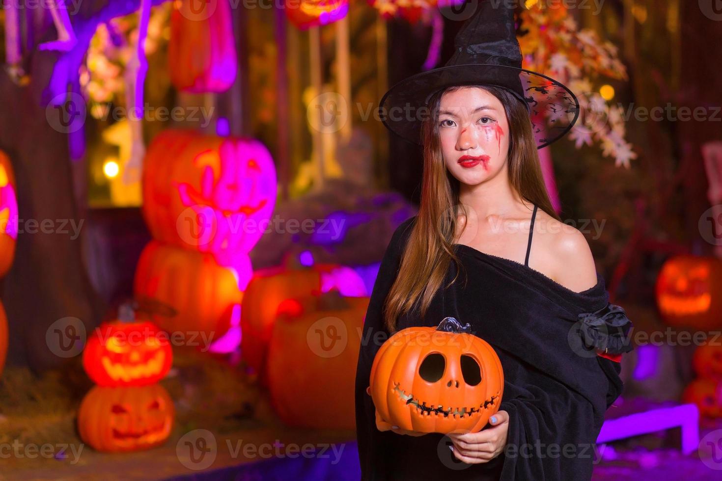 la donna asiatica indossa un cappotto nero fantasma e si trucca come un fantasma di strega mentre guarda la telecamera e tiene in mano una lanterna a testa di zucca di notte in un ambiente esterno su uno sfondo. foto