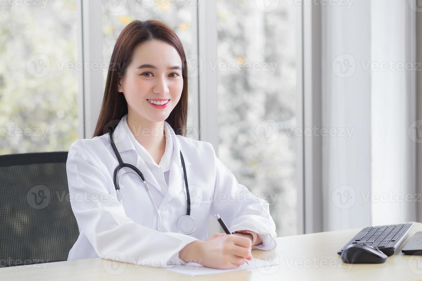 medico donna asiatica professionale indossa cappotto medico e stetoscopio mentre sta scrivendo qualcosa sulla carta e lavorando nella stanza dell'ufficio che sta guardando la telecamera in ospedale. foto